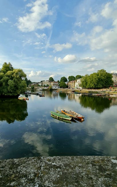 A tranquil riverside scene along the Putney Embankment featuring calm, reflective water with a few small, colorful boats moored along the bank. The garden area in the foreground displays well-maintain